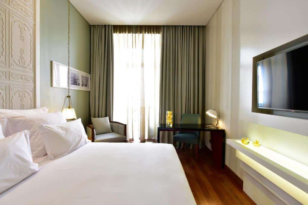 Quarto da Pousada de Lisboa - Small Luxury Hotels Of The World com uma pequena varanda com cortinas, uma mesa com um candeeiro e uma cadeira estofada, um cadeirão estofado, a cama de casal e, em frente, uma televisão