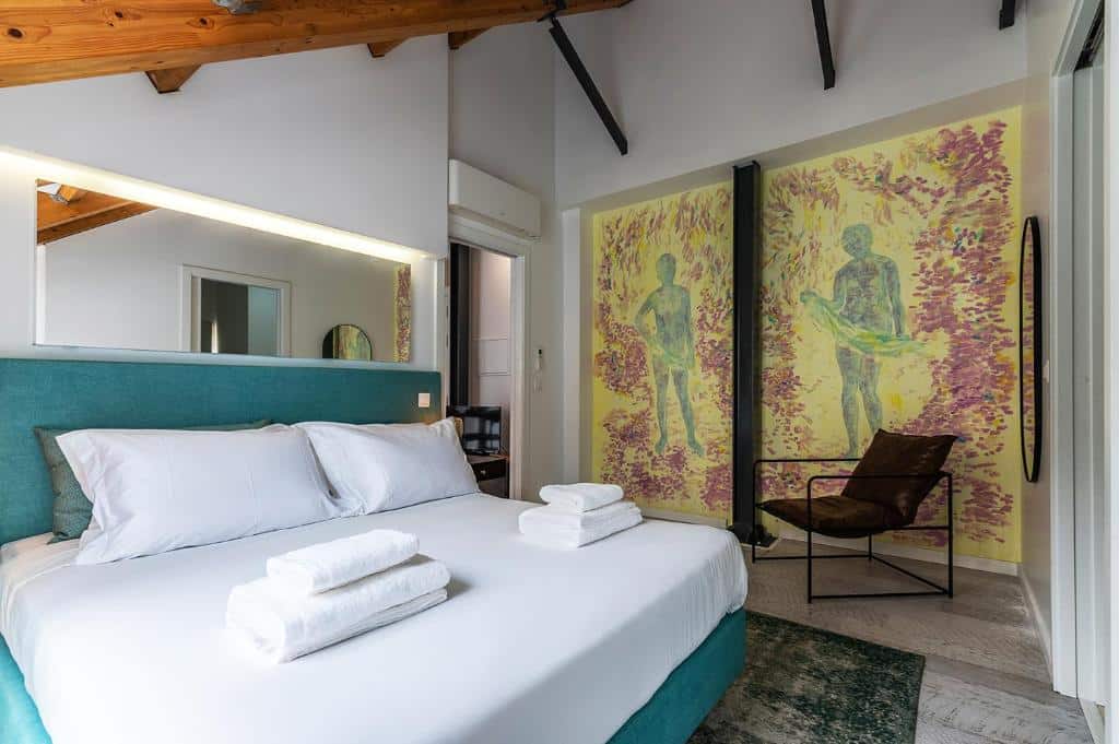 Quarto acolhedor do Portas do Céu com cama azul-petróleo, lençóis brancos e almofadas. Uma obra de arte com duas figuras na parede e uma cadeira a um canto.