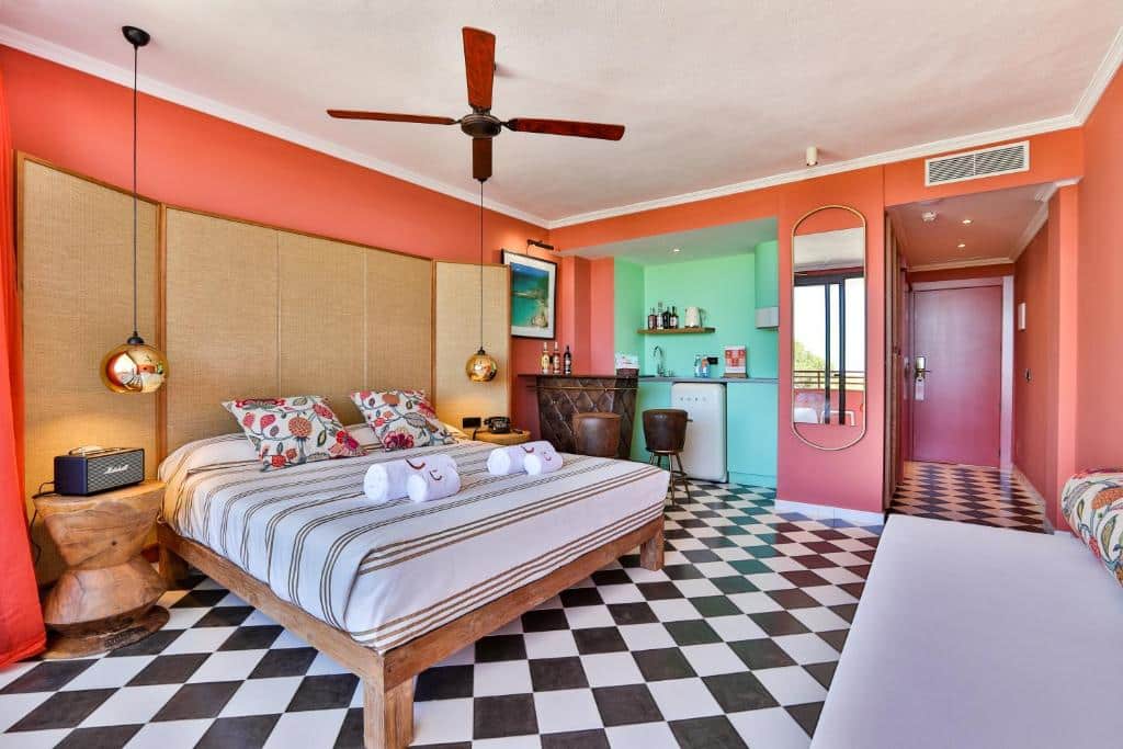 Quarto colorido do Cubanito Ibiza com cama de casal, ventoinha de teto, zona de bar e chão de mosaico xadrez a preto e branco.