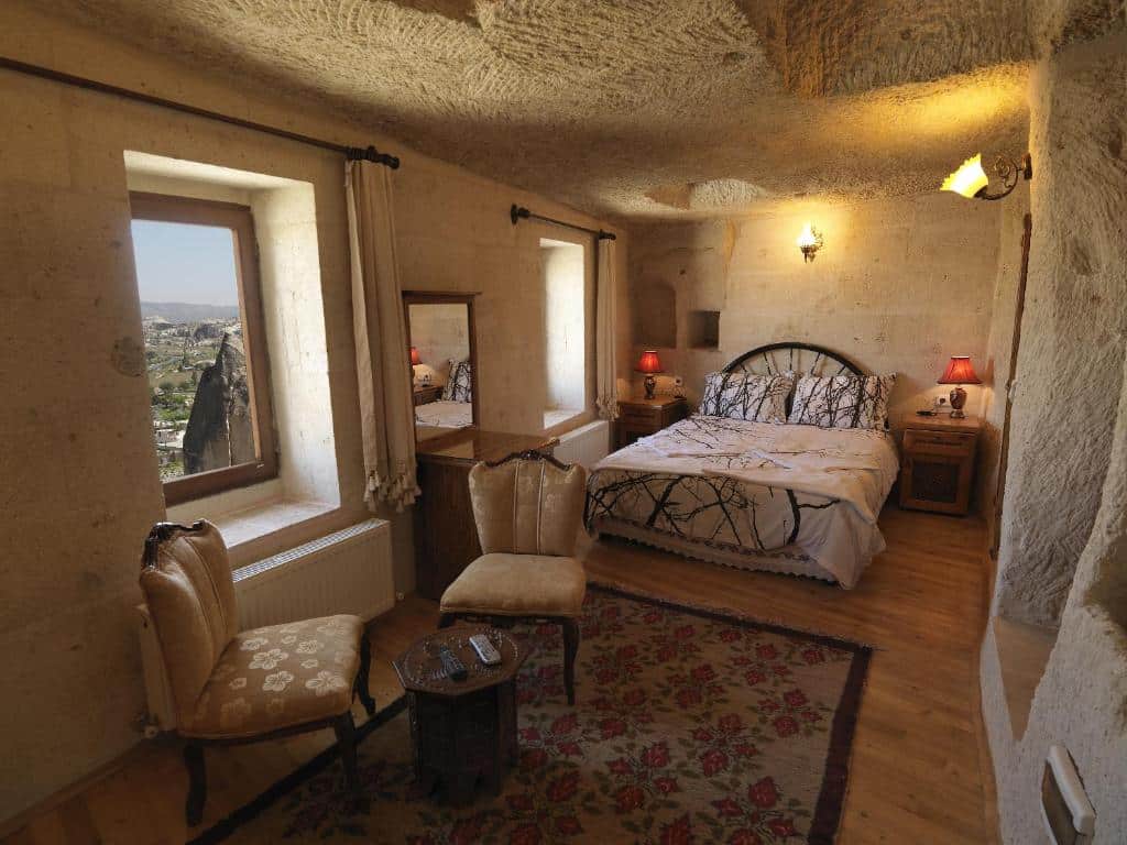 Quarto acolhedor do Arif Cave Hotel, um dos hotéis da Capadócia, com paredes de pedra, cama de casal, duas cadeiras, mesa pequena, janelas, candeeiros e um tapete estampado.