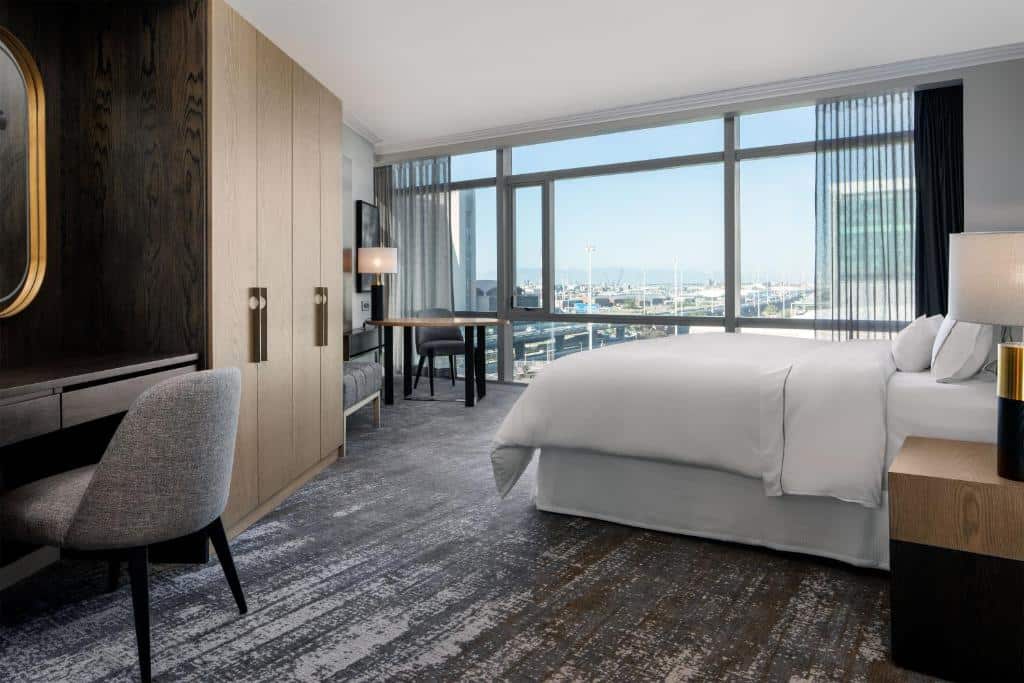 Quarto do The Westin Cape Town com cama grande, secretária, cadeira e janelas do chão ao teto com vista para a cidade e o porto.
