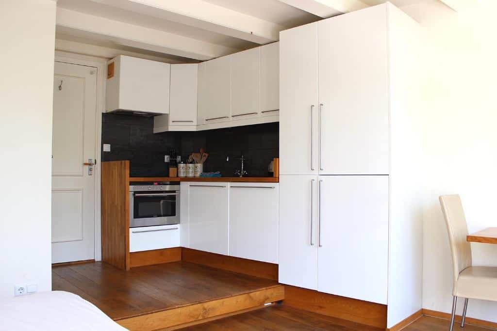 Cozinha moderna do Amsterdam Jewel Canal Apartments, com armários brancos, forno embutido, revestimento preto e pavimento de madeira num espaço pequeno e iluminado.