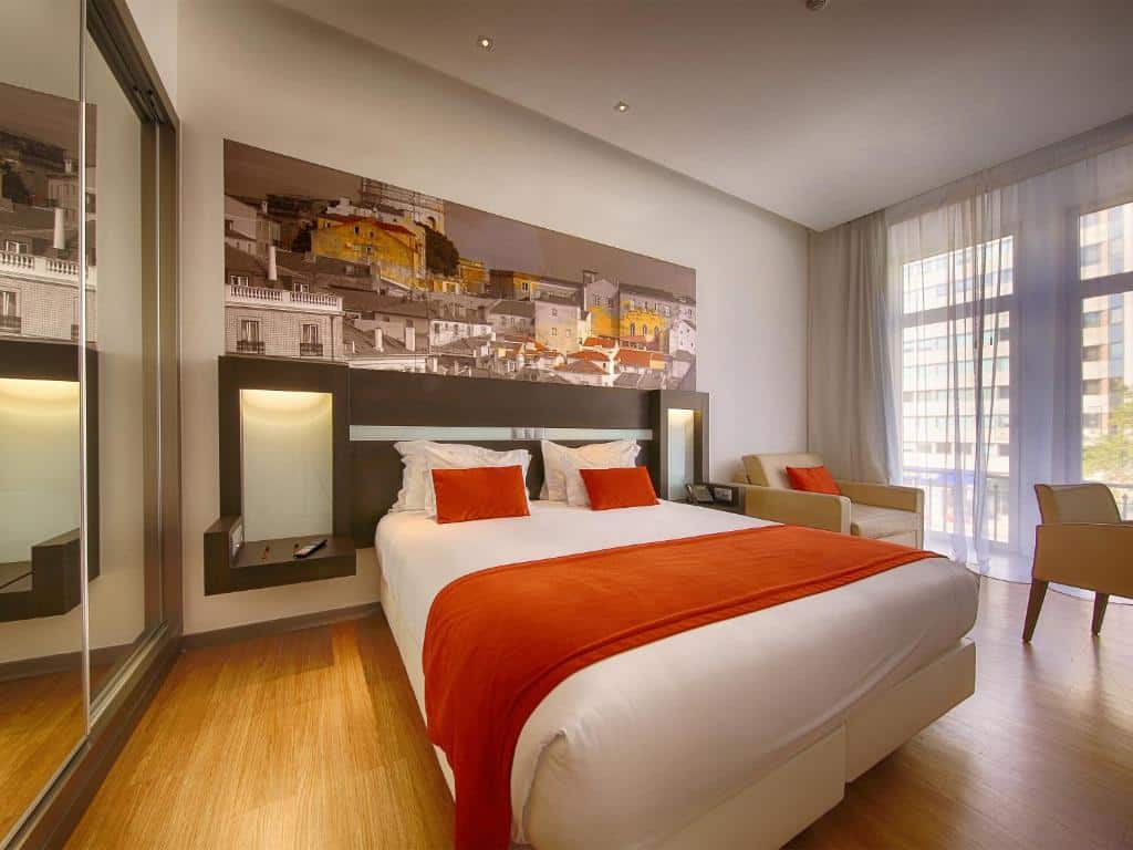 Quarto no Jupiter Lisboa Hotel, com cama de casal ampla, mesa de cabeceira, quadro de Lisboa, poltrona e cortina de voile junto à varanda.