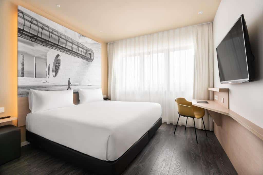 Quarto do Ikonik Lisboa com uma cama de casal, uma janela ampla com cortinas brancas, uma televisão em frente à cama e uma pequena secretária junto à janela, com cadeira