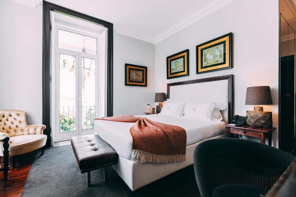 Quarto do Torel Palace Lisbon com uma varanda de vidro, uma cama de casal com almofadas e uma manta, uma cadeira estofada, duas mesinhas de madeira com candeeiros e alguns quadros nas paredes