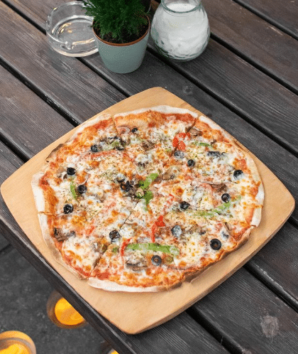 Uma piza inteira do Terraplana coberta com queijo, azeitonas, fatias de pimento vermelho e ervas, numa tábua de madeira sobre uma mesa.