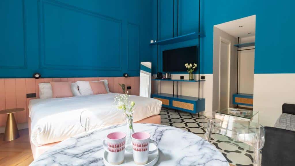 Quarto do Musto Boutique Apartments com paredes azuis, pavimento de ladrilhos preto e branco, cama de lençóis claros, mesa de cabeceira com decoração floral e televisão na parede.