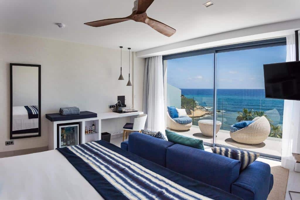 Quarto moderno do Catalonia Royal Ses Estaques com cama, secretária, sofá e varanda com vista para o mar e duas espreguiçadeiras.