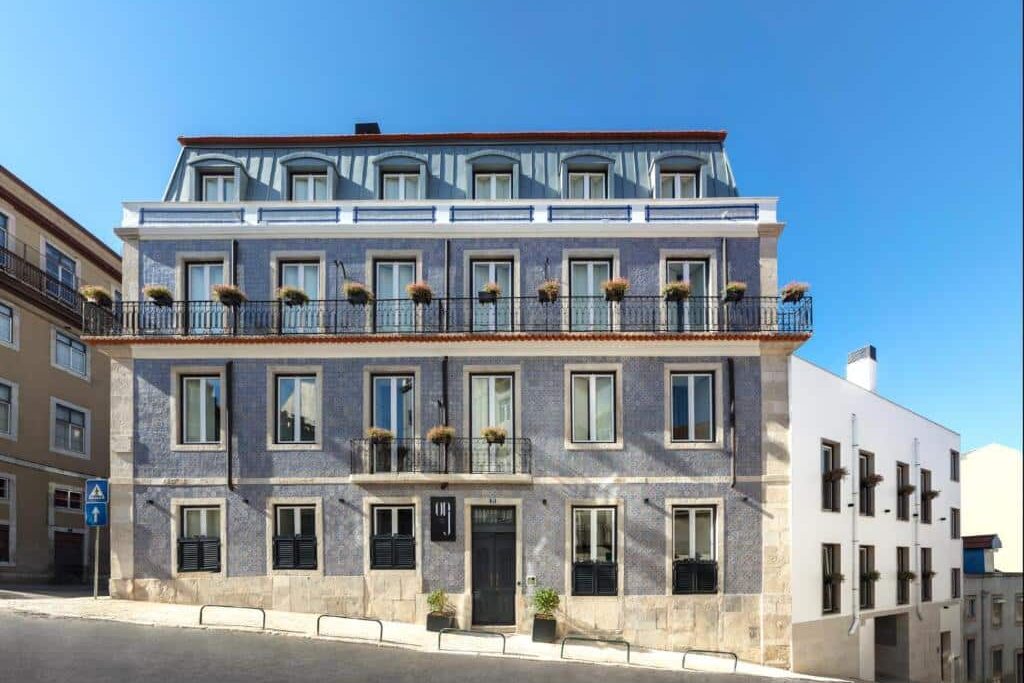 Prédio de quatro andares do OnJ S. Lázaro Lisboa com fachada de azulejos azuis, várias janelas e varandas com vasos de plantas, numa rua ligeiramente inclinada.