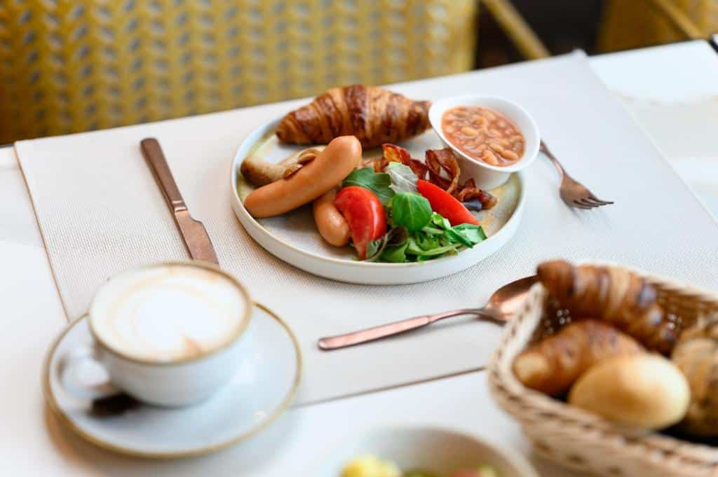 Um prato de pequeno-almoço do Hotel Vision Budapest com salsichas, bacon, croissant, feijão, salada, uma chávena de café e pão num cesto.