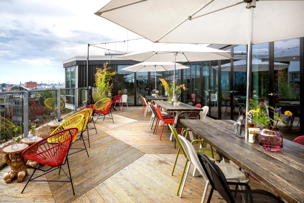 Pátio exterior na cobertura do Zoku Amsterdam, com cadeiras coloridas, mesas compridas de madeira, guarda-sóis e vasos de plantas junto a janelas de vidro.