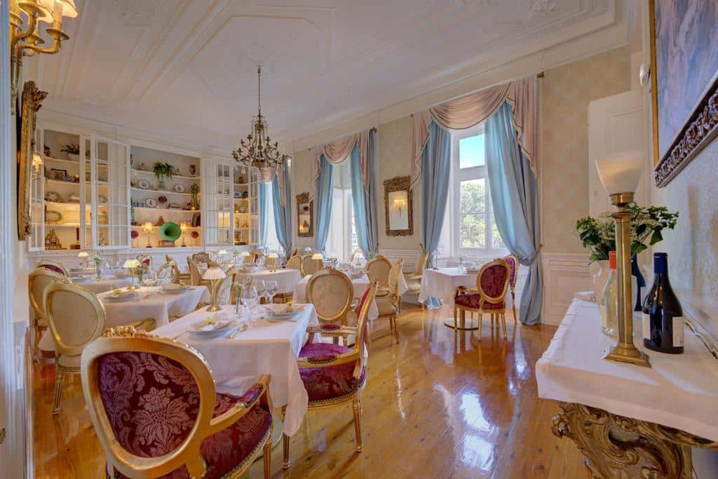Sala de refeições do Palácio das Especiarias, com decoração luxuosa, janelas de cortinas brancas e azul-claro, mesas e cadeiras estofadas em vermelho e dourado e um lustre dourado