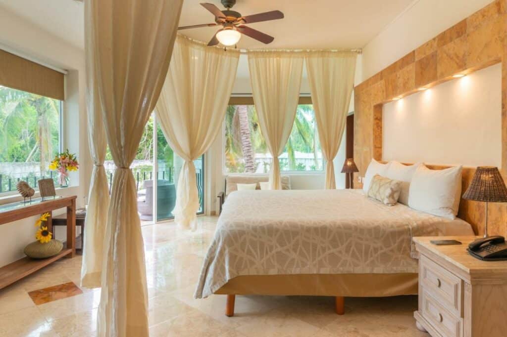 Quarto no Villa Palmeras com cortinas em redor da cama a imitar um bangaló, espaço amplo com varanda, cama de casal, ventoinha e vasos de flores, em tons de bege e branco