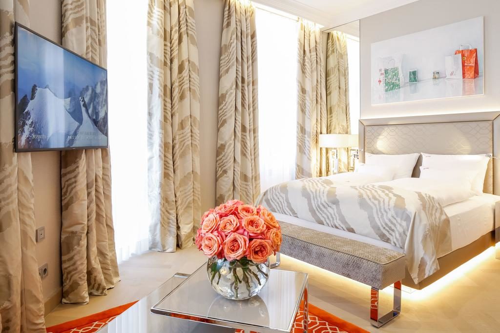 Quarto no Boutiquehotel Das Tyrol, com cama de casal, uma TV na parede, janelas grandes e uma mesa com um vaso de rosas.