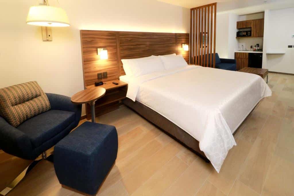 Quarto do Holiday Inn Express & Suites com cama de casal, poltrona azul e mesa de cabeceira com candeeiro.