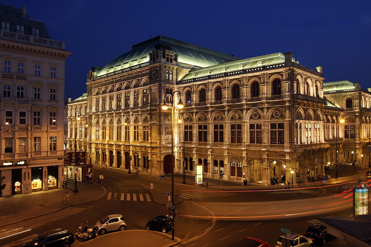 Ópera de Viena à noite, um edifício histórico e ornamentado com janelas em arco e telhado verde, iluminado por candeeiros.