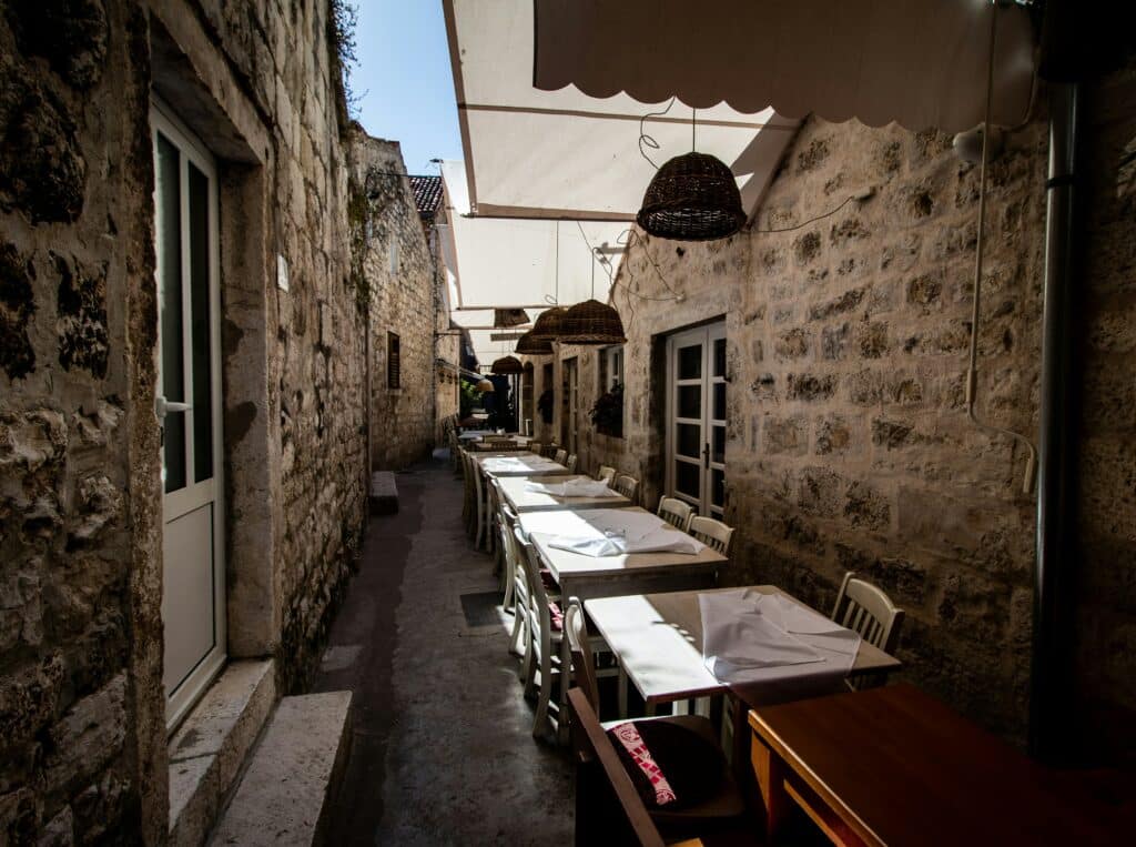 Beco estreito ao ar livre em Hvar, com paredes de pedra, ladeado por mesas e cadeiras brancas sob toldos, que conduz a uma entrada ao fundo.
