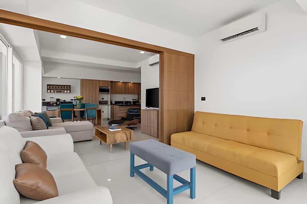Sala de estar moderna do Monyxbnb at Zoho Skies com sofás brancos e amarelos, um banco azul e vista aberta para a cozinha e a zona de refeições.