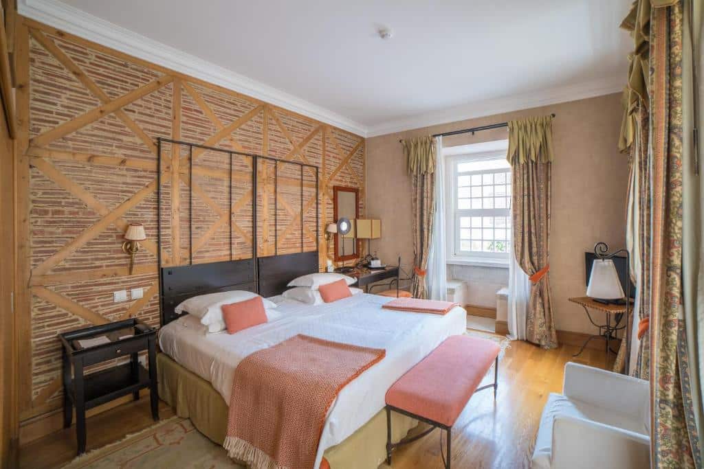Quarto do Solar do Castelo em estilo de época, com uma janela de cortinas, uma pequena mesa com cadeira, uma mesinha com televisão, a cama de casal ao centro e uma poltrona estofada em frente