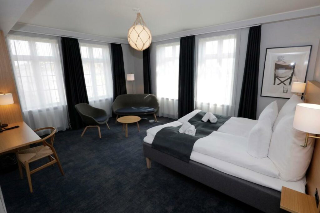 Quarto do Hotel Bethel com duas camas juntas a formar uma cama de casal, cadeiras, várias janelas com cortinados e uma mesa.