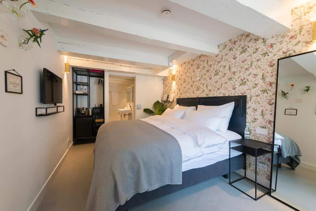 Cama bem arrumada com roupa de cama branca num quarto moderno do Milkhouse Luxury Stay Amsterdam, com papel de parede floral e uma TV na parede.