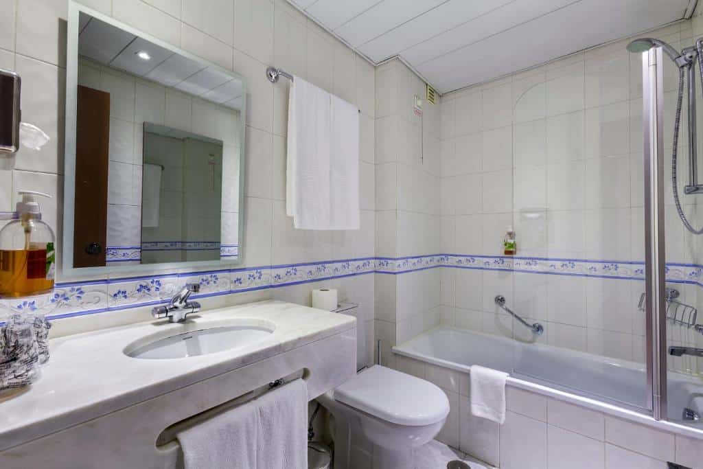 Casa de banho com azulejos brancos, lavatório, espelho, sanita, toalha e banheira com resguardo de vidro e acabamento floral azul, num quarto da Avenida de Fátima.