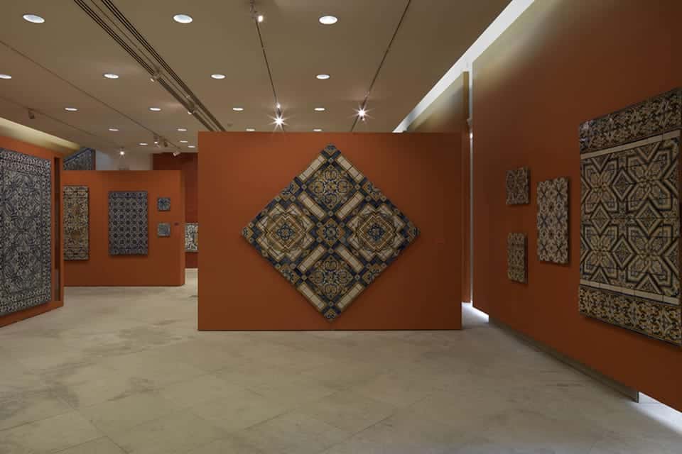 Sala de exposição no Museu Nacional do Azulejo, em Lisboa, com vários azulejos de padrões complexos em paredes cor de laranja.