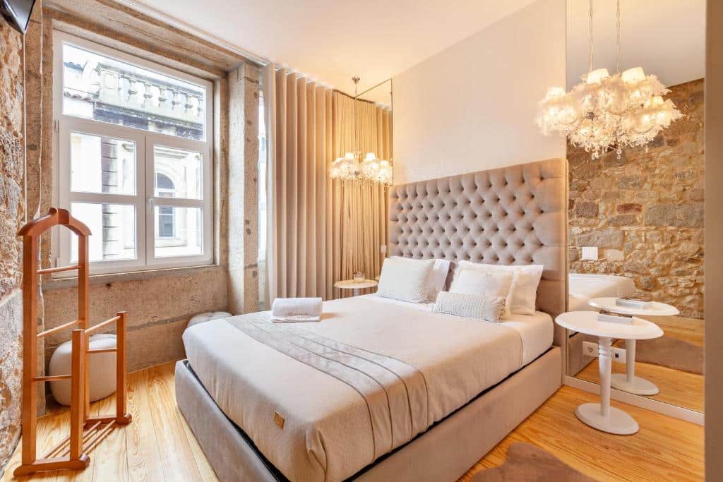 Cama bem feita num quarto luminoso do Berço de Mordomias, com paredes de pedra, janela, espelho e candeeiros modernos.