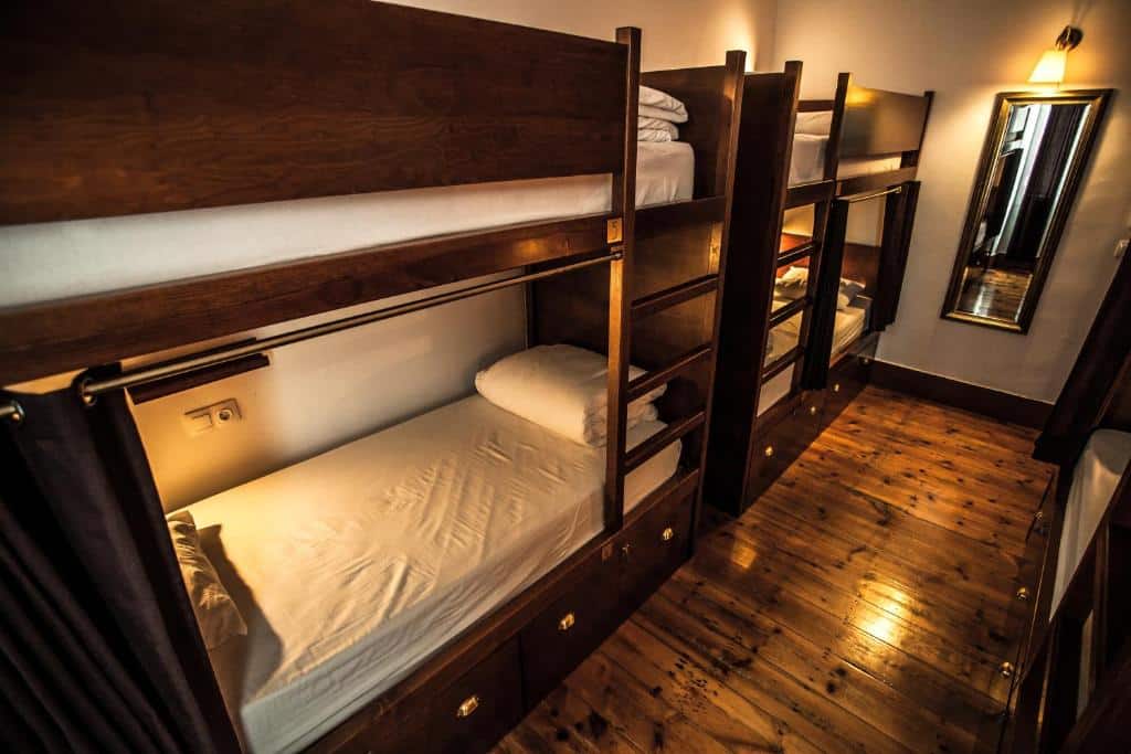 Quarto estreito com beliches de madeira, roupa de cama branca, chão de madeira e um espelho na parede ao fundo, no Home Lisbon Hostel.
