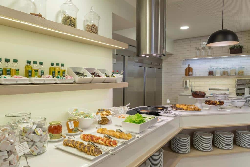 Balcão de buffet com bolos, saladas, condimentos, pratos e garrafas de azeite dispostos de forma organizada sob boa iluminação, no Hotel Cinquentenário & Conference Center.