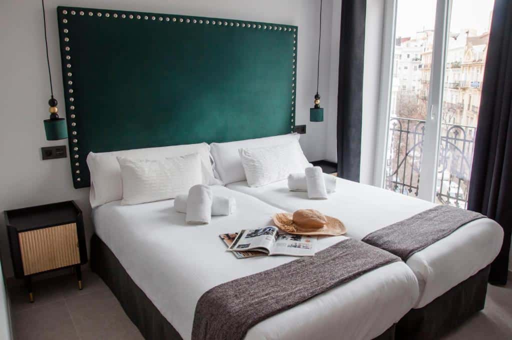 Quarto do Valencia Apartamentos Goya com cabeceira verde-musgo e apontamentos em preto, cama branca com lençóis e toalhas, um livro e um chapéu de palha sobre a cama e janela com vista exterior.