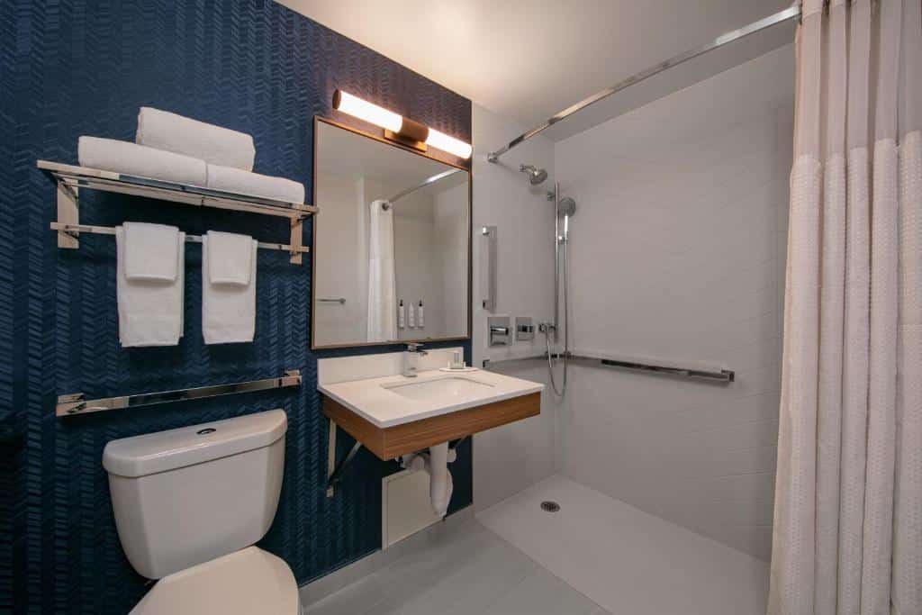 Casa de banho do Fairfield Inn by Marriott JFK Airport com lavatório branco e torneira moderna à esquerda e um espelho retangular com luzes embutidas. À direita, a sanita e, ao fundo, uma cabine de duche com cortinado branco.