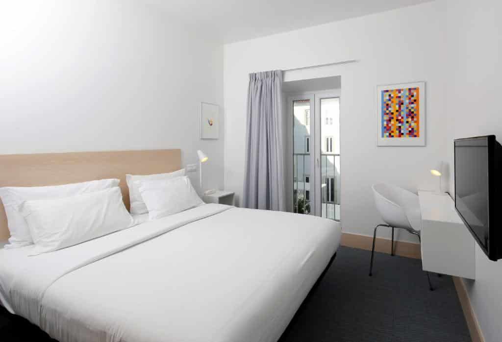 Quarto moderno no Hotel Convento do Salvador com cama de casal, secretária, cadeira, televisão na parede e uma janela com cortinas cinzentas.