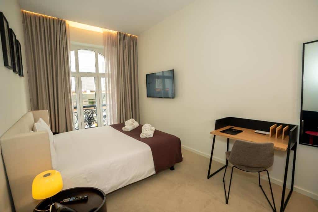 Quarto do Golden Lisbon Hotel com uma pequena varanda com cortinas, uma cama de casal com televisão em frente, uma secretária com cadeira e um espelho