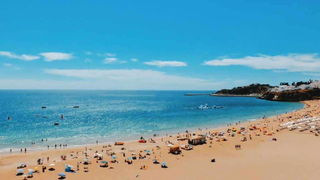 Praia de Albufeira, no Algarve, de areia repleta de pessoas, chapéus de sol e mar azul sob um céu limpo; falésias e edifícios visíveis ao longe.