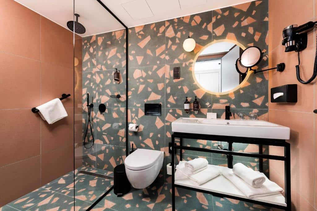 Casa de banho do Three Corners Downtown Hotel com pavimento em terrazzo, chuveiro walk-in, sanita suspensa, espelho redondo e lavatório com toalhas por baixo.