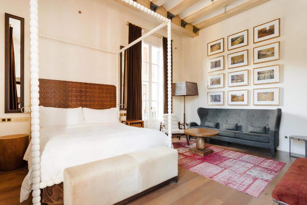 Quarto do Sonder DO Plaça Reial, com cama de casal de dossel branco, mesas de cabeceira de ambos os lados e uma zona de estar com sofá, mesa de centro e quadros na parede.