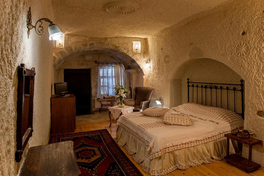 Quarto acolhedor do Aydinli Cave Hotel, um dos hotéis da Capadócia, com paredes de pedra, cama em ferro forjado, tapete, poltrona, televisão e uma janela com cortinados transparentes.