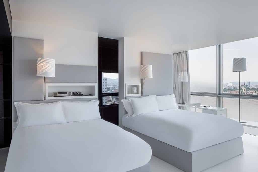 Quarto no SO/ Vienna, com duas camas de casal, roupa de cama branca, decoração moderna, janelas grandes e vista para a cidade e para a água.