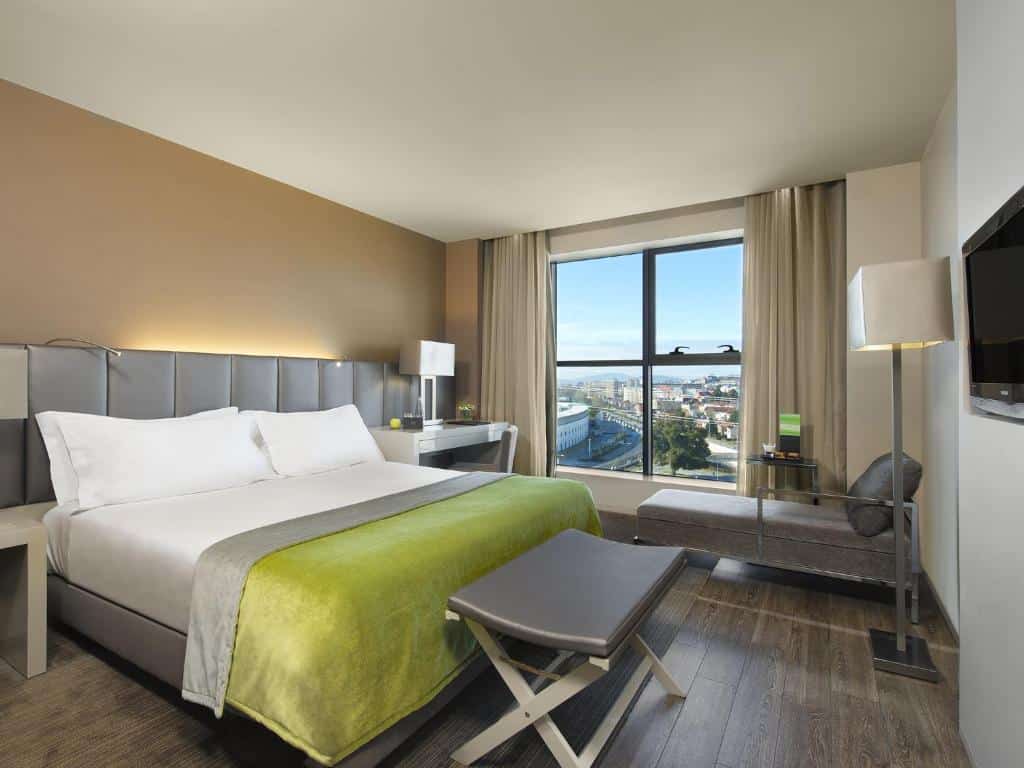 Quarto moderno do Meliá Braga Hotel & Spa com cama de casal, colcha verde, secretária, candeeiros e janela ampla com vista para a cidade.