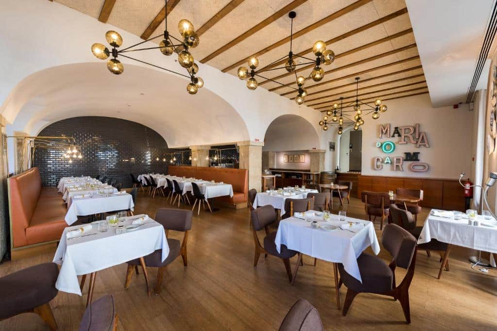 Sala de refeições do Lisboa Carmo Hotel com candeeiros suspensos, mesas de madeira com cadeiras estofadas e sofás em tons de caramelo.