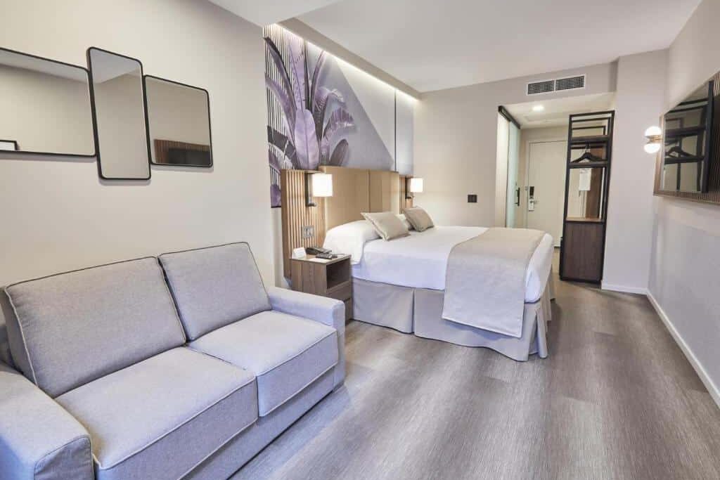 No quarto do Riu Plaza Manhattan Times Square há uma cama de casal ao centro, com cabeceira estofada. À esquerda, uma secretária com espelho por cima e uma TV na parede; à direita, uma poltrona e uma mesa de cabeceira com candeeiro. A janela ao fundo tem vista para a cidade.