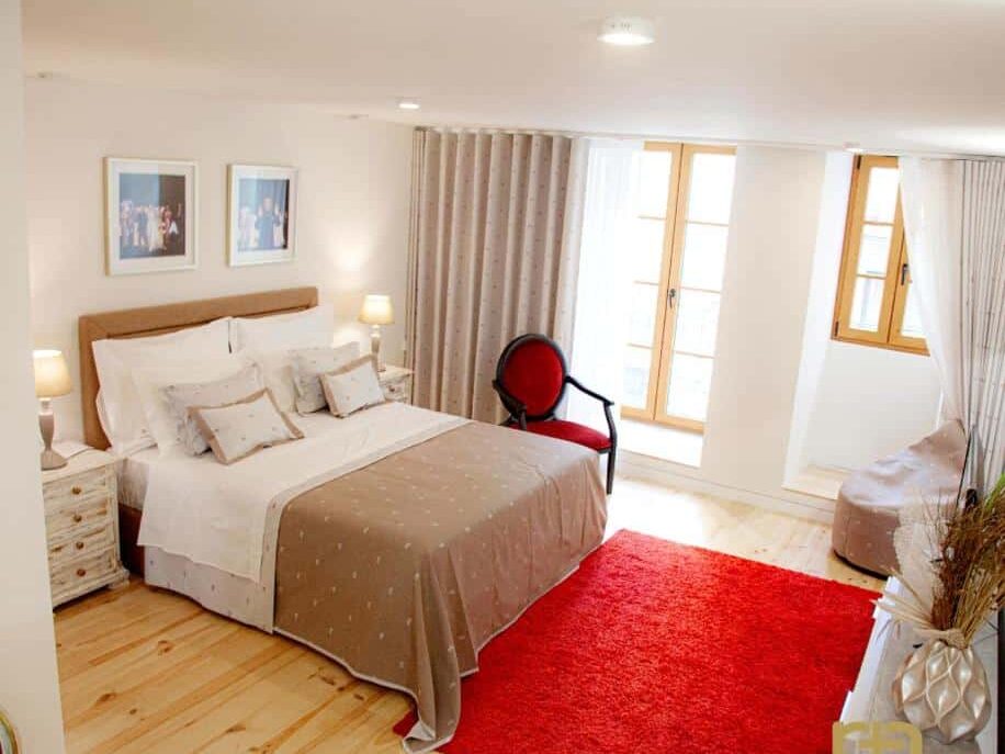 Quarto acolhedor do TM Luxury Apartments Lisbon com cama de casal, alcatifa vermelha, chão de madeira e duas janelas com cortinas. Dois candeeiros em mesas de cabeceira.