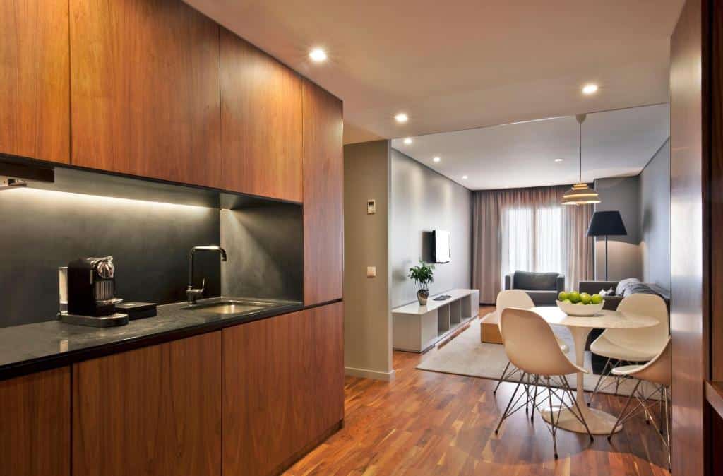Cozinha compacta do Altis Prime Hotel com armários, uma máquina de café e um lava-loiça; seguindo pela mesma parede há um pequeno corredor e, depois, uma sala de estar com uma mesa redonda com quatro lugares, uma televisão, um sofá, uma janela com cortinas e um candeeiro de pé ao lado de uma poltrona estofada