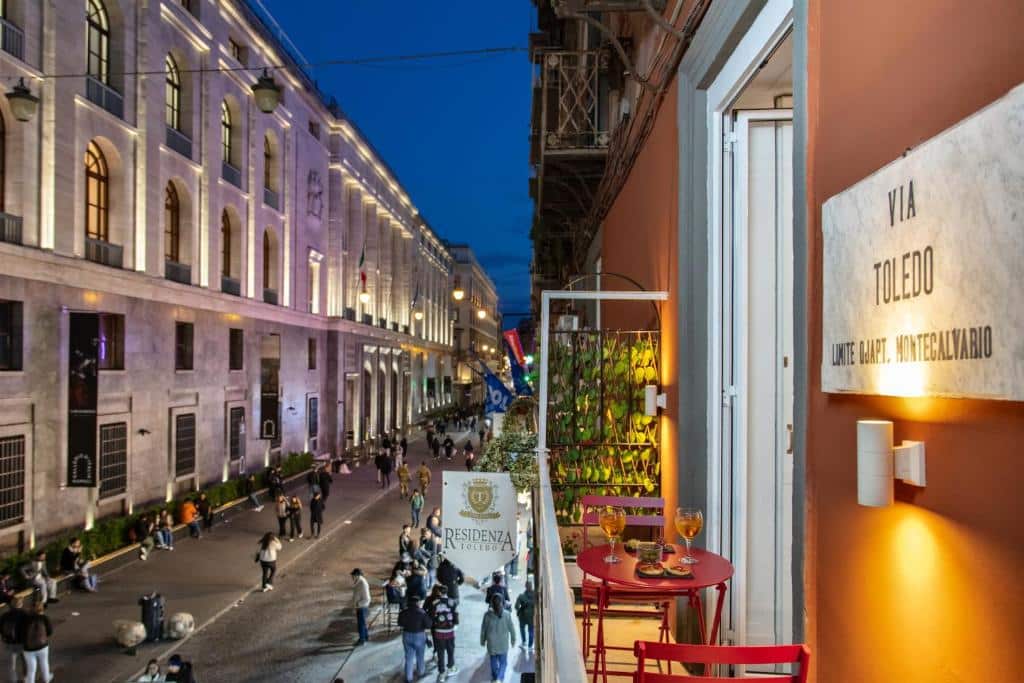 Rua iluminada com edifícios históricos e um restaurante com mesas e cadeiras vermelhas no passeio, pessoas a caminhar à noite, junto ao Residenza Toledo.