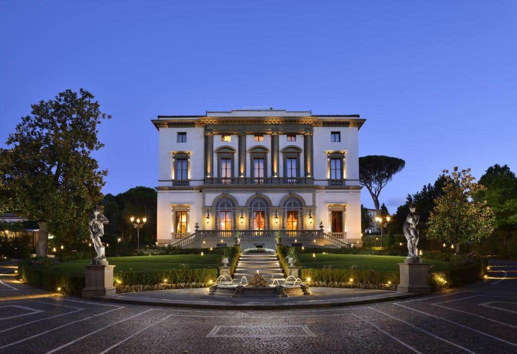 Edifício clássico iluminado numa noite de um dos hotéis na Toscana, o Villa Cora, com um chafariz à frente, duas estátuas ao lado, jardim verde de relva baixa e uma escadaria de acesso ao edifício