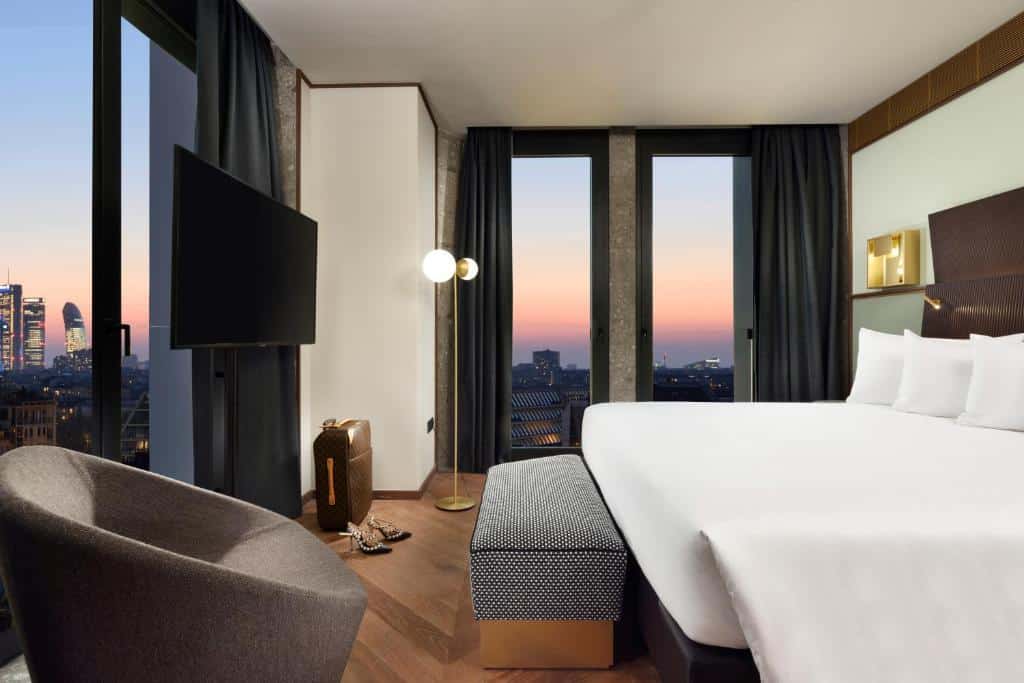 Quarto do Milano Verticale | UNA Esperienze com janelas panorâmicas, uma cama de casal, um candeeiro de pé, uma poltrona e uma televisão em frente à cama