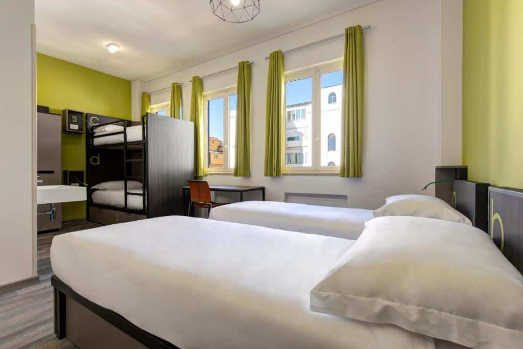 quarto do The RomeHello, um dos hostels em Roma, com um beliche e duas camas de solteiro com colchas brancas e três janelas quadradas com cortinas, num ambiente arejado