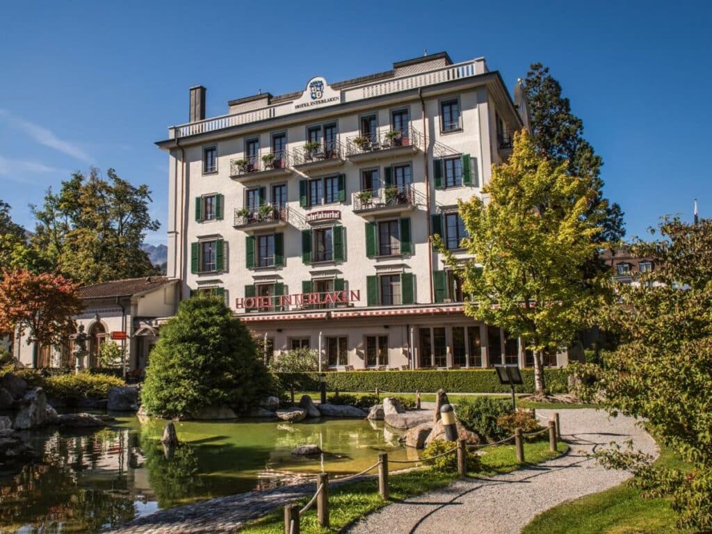 Fachada do Hotel Interlaken durante o dia, com jardim e lago em frente.