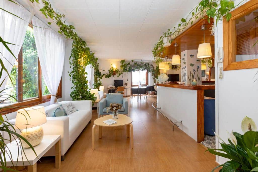 Sala de estar bem iluminada no Hostal Manolita, com mobiliário branco, detalhes em madeira, muitas plantas e grandes janelas.